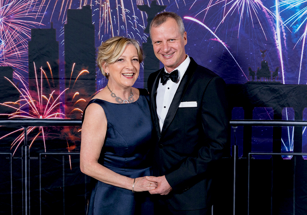 Webseite_GF_neuesFoto Kopie links Frau Sabine Fliegner in blauem Ballkleid, rechts Herr Ulrich Fliegner im schwarzen Smoking, Hintergrund Brandenburger Tor mit Silvesterfeuerwerk